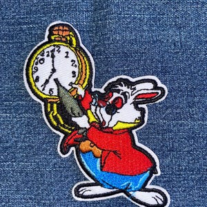 May include: Embroidered patch featuring the White Rabbit from Alice in Wonderland, holding a pocket watch. The rabbit is depicted in a red jacket, blue pants, and white fur, with a yellow and black watch. The patch has a white border.