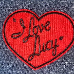 May include: A red heart-shaped patch with the phrase "I Love Lucy" embroidered in black script. The patch has a black border and is set against a blue denim background.