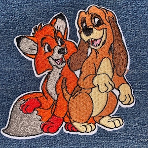 May include: Embroidered patch featuring the characters Tod the fox and Copper the hound dog from the animated film "The Fox and the Hound." The patch has a white border and is set against a blue denim background. The fox is orange and the dog is brown.