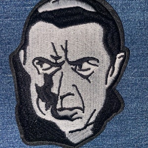 May include: Embroidered patch featuring a black and gray portrait of a stern-faced man with slicked-back hair. The patch has a black border and is likely for application on clothing or accessories. The design is detailed and stylized.