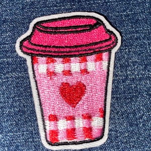 May include: An embroidered patch shaped like a coffee cup with a bright pink lid. The cup features a light pink body with red and white checkered patterns at the top and bottom, and a solid red heart centered on the front of the cup.
