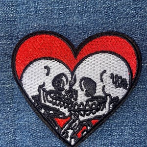 May include: A heart-shaped embroidered patch featuring two white skulls facing each other in profile. The skulls are set against a vibrant red background within the heart, and the entire design is outlined with a thick black border.