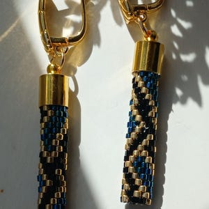 May include: A pair of drop earrings featuring cylindrical beaded dangles in a repeating pattern of metallic gold and dark blue glass beads. Each earring is attached to a polished gold-tone hinged lever-back finding with a smooth, rounded finish.