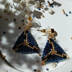 Pu&ograve; includere: Un paio di orecchini a lobo triangolari realizzati con piccole perline blu scuro e oro metallizzato. Gli orecchini presentano un motivo geometrico con accenti dorati e includono perni in metallo dorato con chiusure in plastica trasparente.