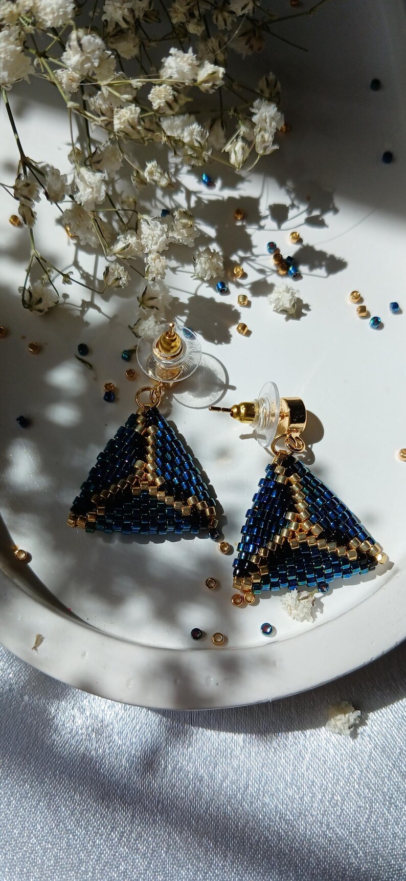 Pu&ograve; includere: Un paio di orecchini a lobo triangolari realizzati con piccole perline blu scuro e oro metallizzato. Gli orecchini presentano un motivo geometrico con accenti dorati e includono perni in metallo dorato con chiusure in plastica trasparente.