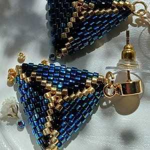 Puede incluir: Un par de pendientes triangulares hechos con pequeñas cuentas de semillas en azul iridiscente y oro metálico. El diseño geométrico presenta un patrón repetitivo de cuentas azules y doradas, con postes de metal dorado y cierres transparentes.