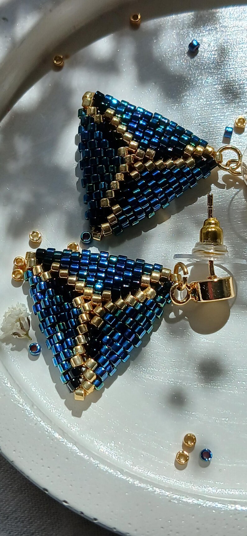 Pu&ograve; includere: Un paio di orecchini triangolari realizzati con piccole perline di conteria blu iridescente e oro metallizzato. Il design geometrico presenta un motivo ripetuto di perline blu e dorate, rifinito con perni in metallo dorato e chiusure trasparenti.
