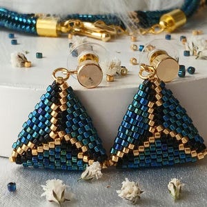 Pu&ograve; includere: Un paio di orecchini triangolari realizzati con piccole perline iridescenti blu, nere e oro metallizzato. Gli orecchini presentano perni in metallo dorato con chiusure in plastica trasparente e sono accompagnati da una collana di perline.
