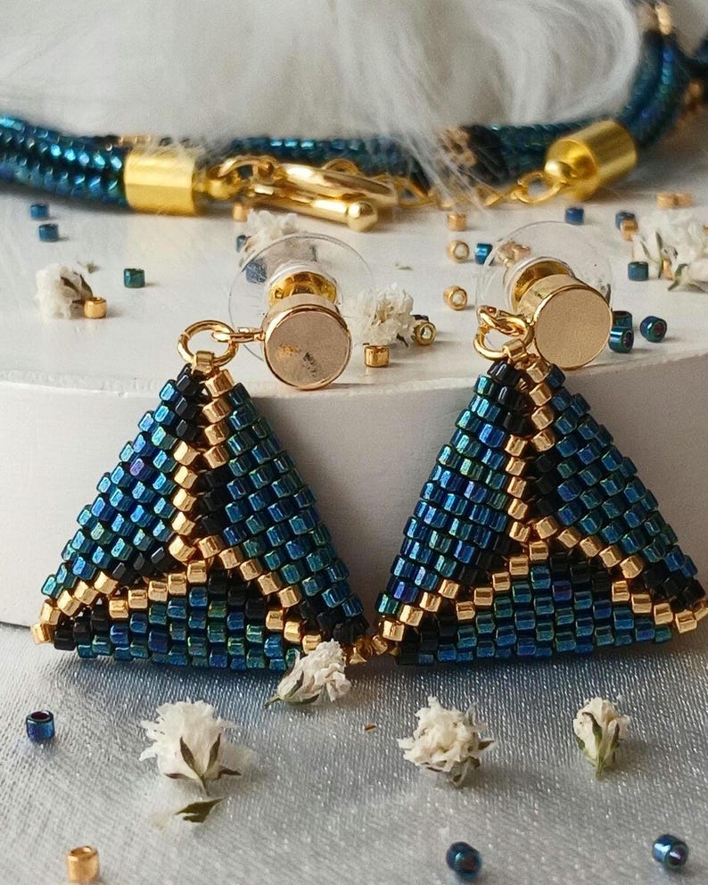 Pu&ograve; includere: Un paio di orecchini triangolari realizzati con piccole perline iridescenti blu, nere e oro metallizzato. Gli orecchini presentano perni in metallo dorato con chiusure in plastica trasparente e sono accompagnati da una collana di perline.