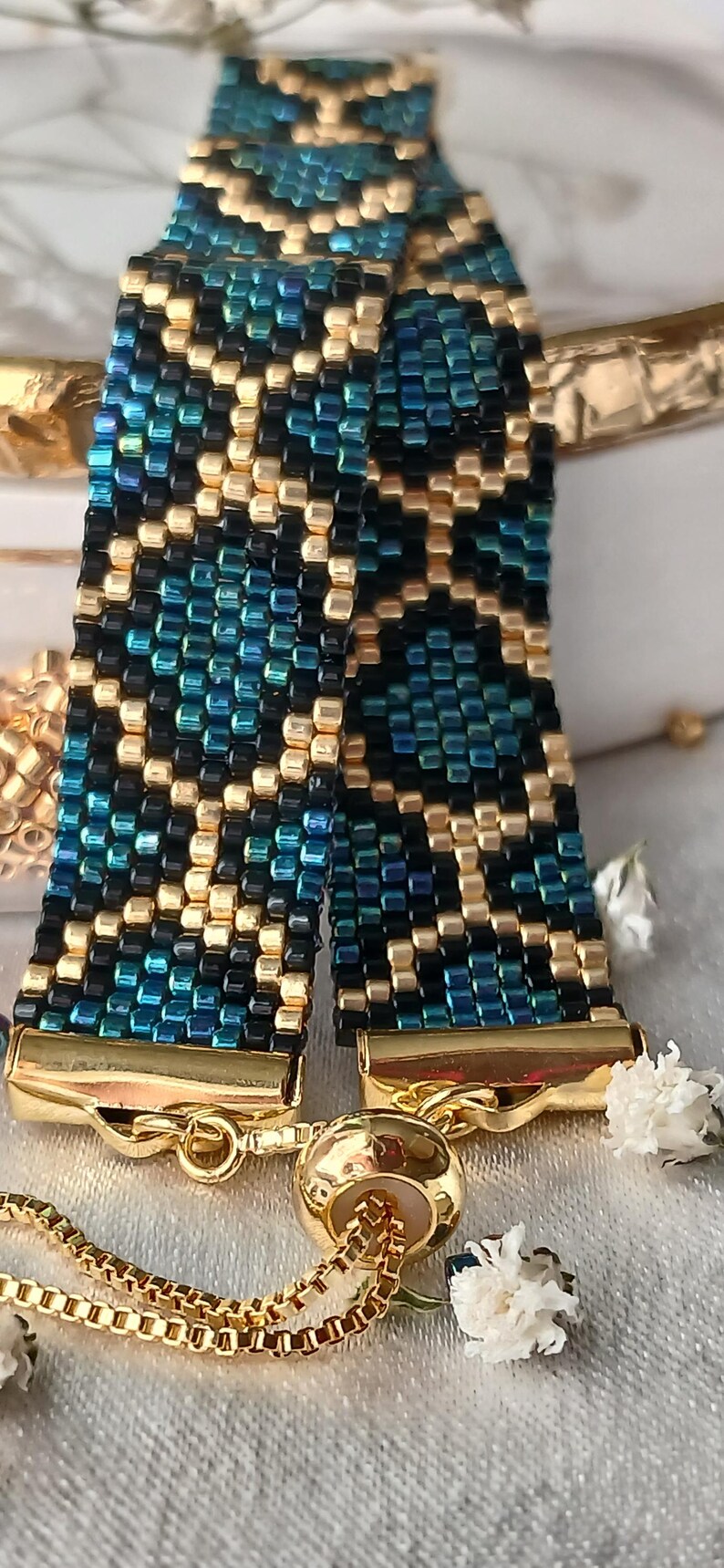 Pu&ograve; includere: Un bracciale in perline intrecciate con un motivo geometrico realizzato in perline di vetro color oro metallizzato, nero profondo e verde acqua iridescente. Presenta terminali in metallo dorato e una chiusura a catena scorrevole con perla.