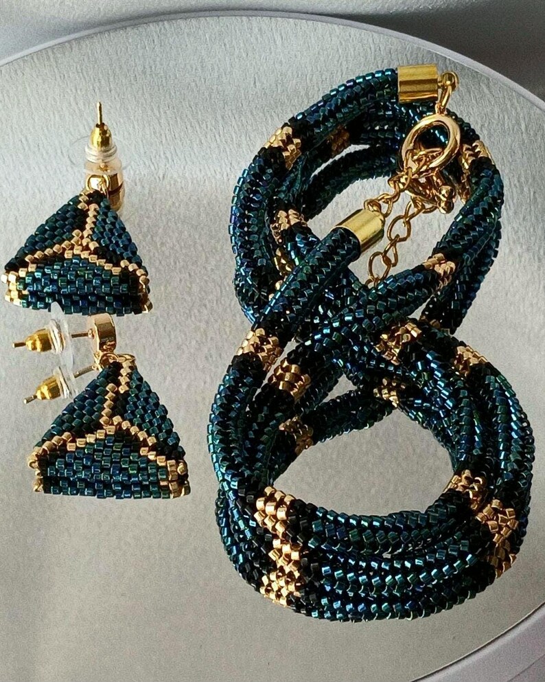 Pu&ograve; includere: Un set di gioielli composto da due orecchini a lobo triangolari e una collana abbinata a corda. I pezzi sono realizzati con piccole perline iridescenti blu petrolio scuro e nere con accenti dorati e componenti in metallo color oro.