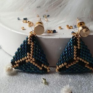 Pu&ograve; includere: Un paio di orecchini triangolari realizzati con piccole perline blu scuro iridescente e oro metallizzato. Gli orecchini presentano un motivo geometrico e sono fissati a perni in metallo dorato con chiusure in plastica trasparente.