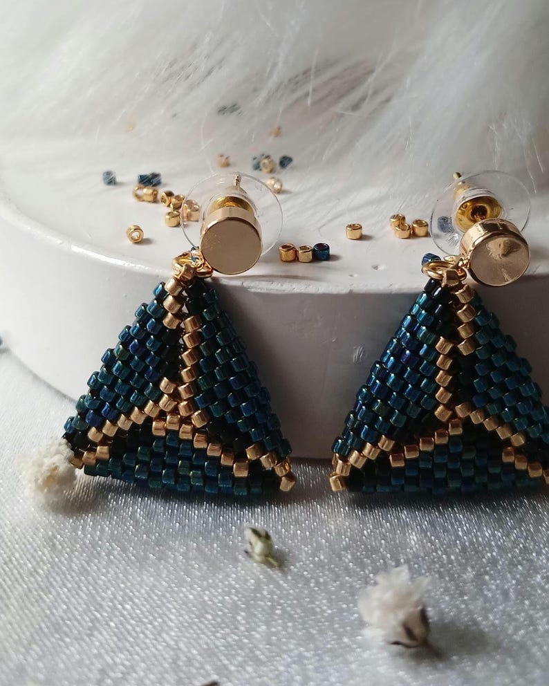 Pu&ograve; includere: Un paio di orecchini triangolari realizzati con piccole perline blu scuro iridescente e oro metallizzato. Gli orecchini presentano un motivo geometrico e sono fissati a perni in metallo dorato con chiusure in plastica trasparente.