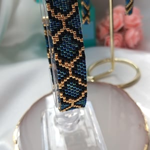 May include: A beaded bracelet and matching drop earrings featuring a geometric pattern of metallic gold, deep blue, and black glass beads. The bracelet is displayed on a clear stand, while the earrings hang from a gold-toned metal jewellery holder.