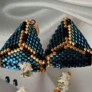 Pu&ograve; includere: Un paio di orecchini triangolari realizzati con piccole perline di conteria blu scuro iridescente e oro metallizzato. Le perline sono disposte in un motivo geometrico, con accenti dorati che formano linee sul lavoro di perline blu brillante.