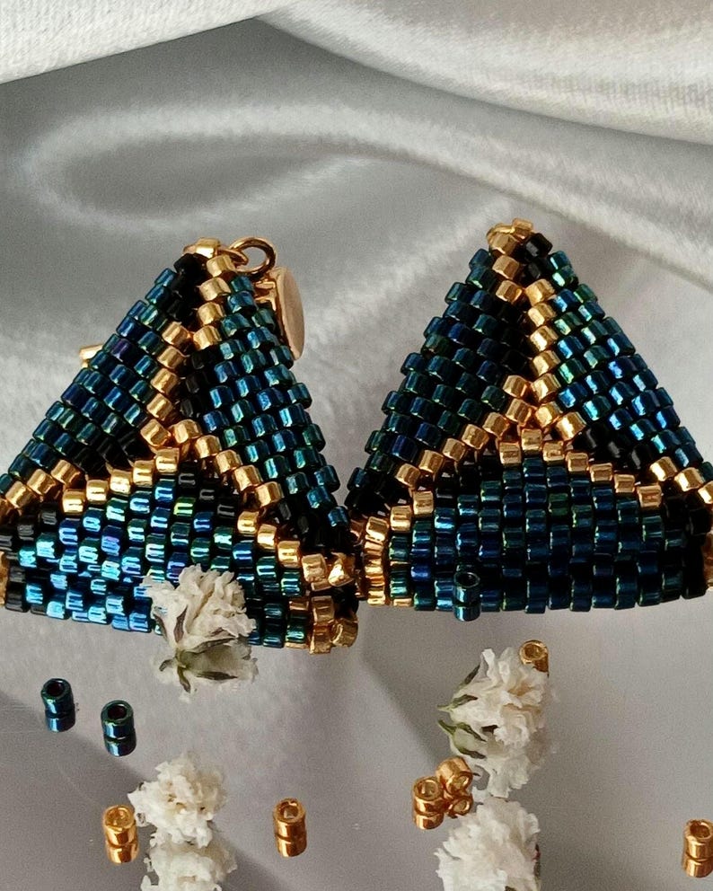 Pu&ograve; includere: Un paio di orecchini triangolari realizzati con piccole perline di conteria blu scuro iridescente e oro metallizzato. Le perline sono disposte in un motivo geometrico, con accenti dorati che formano linee sul lavoro di perline blu brillante.