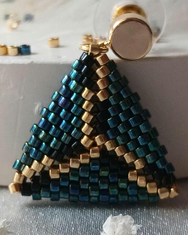 Pu&ograve; includere: Un orecchino triangolare realizzato con piccole perline iridescenti color ottanio, blu scuro e oro metallizzato. La lavorazione geometrica a perline &egrave; fissata a un perno circolare color oro con un inserto centrale liscio rosa pallido.