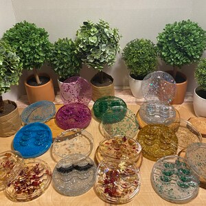 May include: Assortment of 12 round, clear, resin trays with various glitter and decorative accents. The trays have multiple compartments for storing small items like lashes, jewelry, or other accessories.