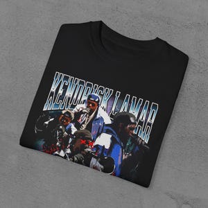 Op de afbeelding: Zwart T-shirt met een vintage-stijl grafische afbeelding van Kendrick Lamar en andere figuren. De tekst "Kendrick Lamar" is gedrukt in een blauw en wit lettertype.