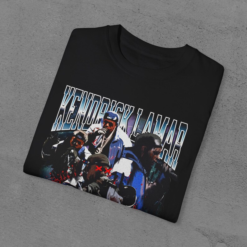 Op de afbeelding: Zwart T-shirt met een vintage-stijl grafische afbeelding van Kendrick Lamar en andere figuren. De tekst "Kendrick Lamar" is gedrukt in een blauw en wit lettertype.