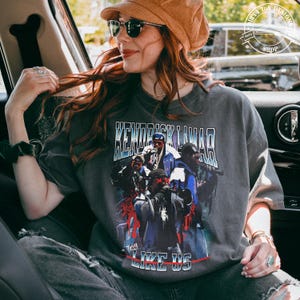 Op de afbeelding: Een grijs T-shirt met een grafische print van Kendrick Lamar en de woorden "To Pimp a Butterfly" en "Like Us".