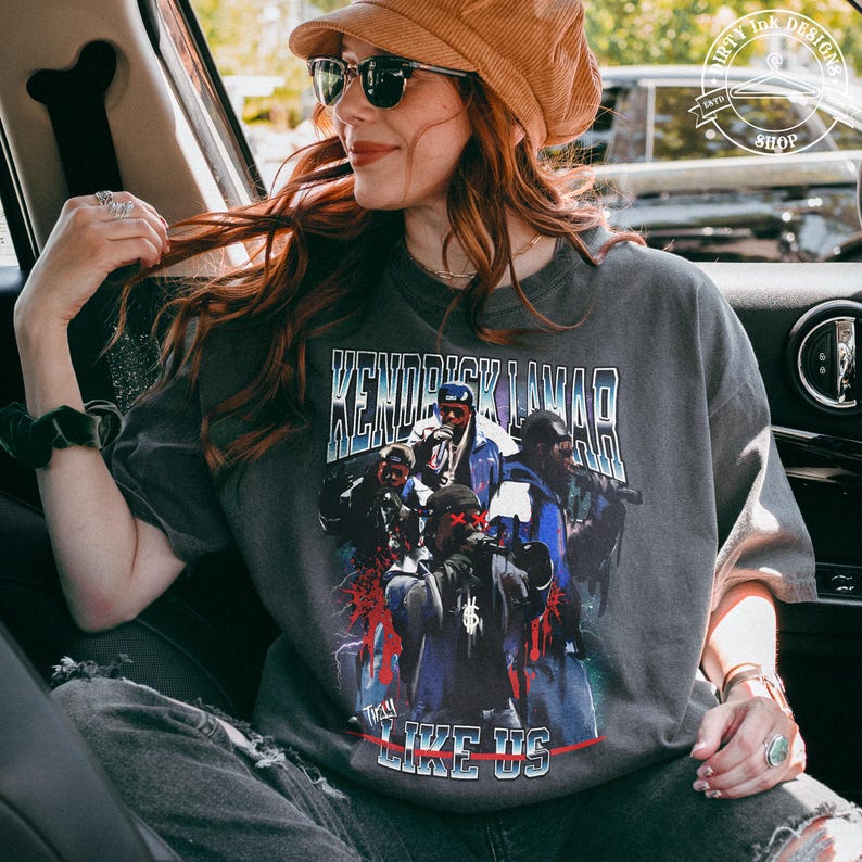 Op de afbeelding: Een grijs T-shirt met een grafische print van Kendrick Lamar en de woorden "To Pimp a Butterfly" en "Like Us".