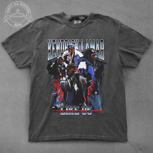 Op de afbeelding: Een donkergrijs T-shirt met een vintage-stijl grafische print van Kendrick Lamar en de woorden "Kendrick Lamar" en "They Like Us".