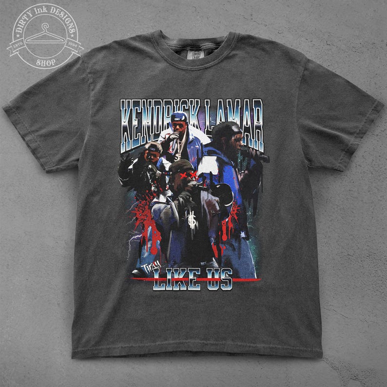 Op de afbeelding: Een donkergrijs T-shirt met een vintage-stijl grafische print van Kendrick Lamar en de woorden "Kendrick Lamar" en "They Like Us".