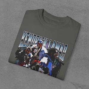 Op de afbeelding: Een grijs T-shirt met een vintage-stijl grafische print van Kendrick Lamar en andere figuren. De tekst "Kendrick Lamar" is gedrukt in een vet, blauw lettertype.