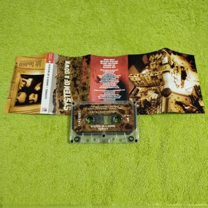 May include: A cassette tape with a clear plastic case. The tape is labeled "System of a Down" and "Toxicity". The tape is part of a larger image that includes a CD case and a booklet with the band's name and album title.