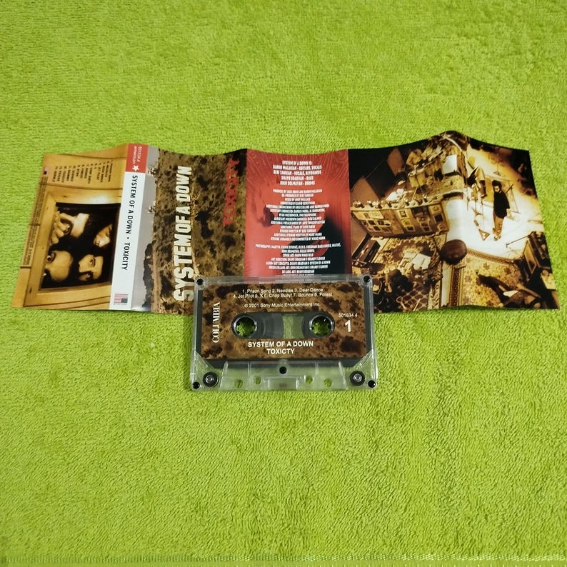 May include: A cassette tape with a clear plastic case. The tape is labeled "System of a Down" and "Toxicity". The tape is part of a larger image that includes a CD case and a booklet with the band's name and album title.
