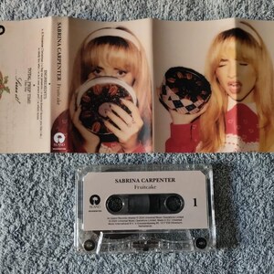 May include: A cassette tape with a white label that reads "Sabrina Carpenter Fruitcake 1". The cassette tape is in a clear plastic case. The background is a close-up of a fabric with a textured pattern.