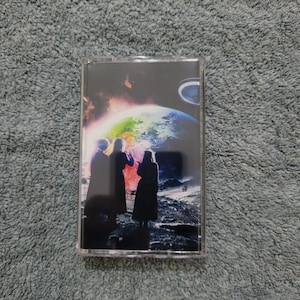 Puede incluir: Una cinta de casete con una imagen colorida de tres figuras de pie sobre una superficie lunar, mirando una Tierra brillante en el fondo.