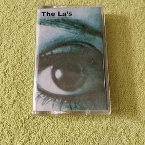 May include: A cassette tape of "The La's" album, featuring an eye on the cover. The image is in shades of teal and green. The cassette is in a clear plastic case, resting on a textured green surface.