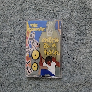 May include: A cassette tape with a yellow, blue, and green design. The tape features a cartoon character holding a large rock with the text "Faith is a Rebel" written on it. The tape is titled "The Alchemist".