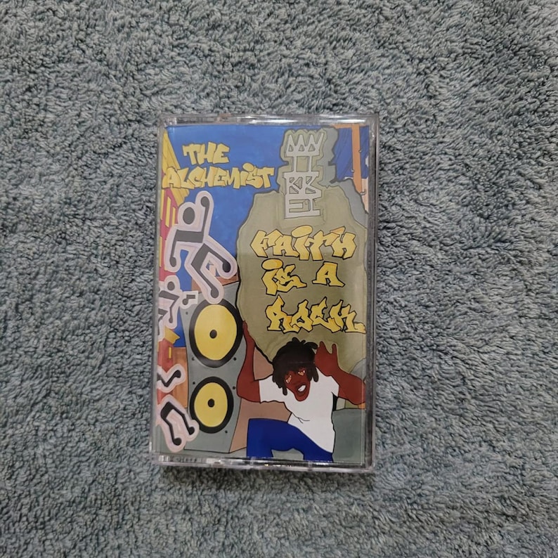May include: A cassette tape with a yellow, blue, and green design. The tape features a cartoon character holding a large rock with the text "Faith is a Rebel" written on it. The tape is titled "The Alchemist".