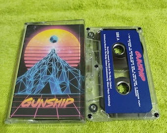 May include: A clear cassette tape case with a blue label that reads "GUNSHIP" and lists the song titles "The Mountain", "Revel in Your", "Tech Nor", and "Shroow Fury". The cassette tape is in a clear plastic case with a black and blue design.