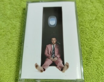 May include: A cassette tape case with a white background and a pink-suited man sitting in a doorway with a blue sky visible through a window. The man is wearing a pink suit, a white shirt, and a tie. The text "Circles" is printed on the case.