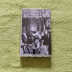 May include: A cassette tape with the word "singles" in white lettering. The cover features a black and white photo of people on a bench and kissing. The cassette is in a clear plastic case.