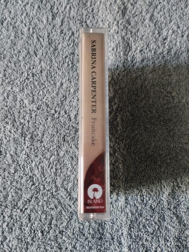 May include: A cassette tape in a clear plastic case. The tape is labelled "Sabrina Carpenter Fruitcake" with the Island Records logo.