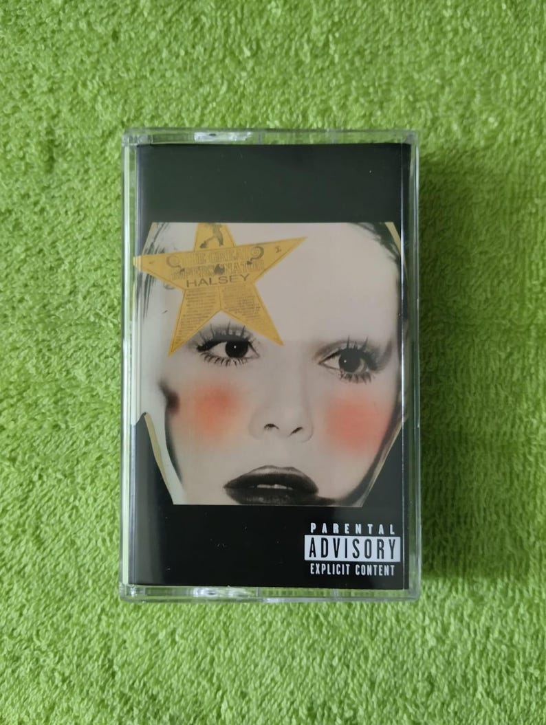 May include: A cassette tape with a black case. The cover features a close-up of a face with a star over one eye. The face has red blush and dark lipstick. The text "PARENTAL ADVISORY EXPLICIT CONTENT" is at the bottom.