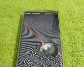 May include: A cassette tape featuring the album "Currents" by Tame Impala. The cover art has a black and white striped background with a pink line and a gray sphere. The text "TAME IMPALA" and "CURRENTS" are in yellow.