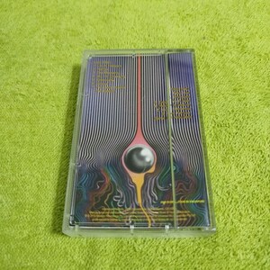 May include: A clear cassette tape with a vibrant, abstract design. The artwork features a central, vertical line with a circular element and flowing, wavy lines in shades of purple, yellow, and blue. Text is visible on the cassette.