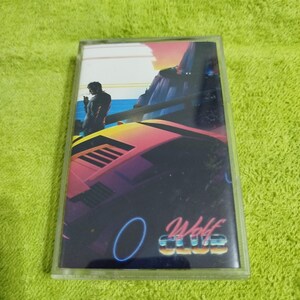 May include: A vintage cassette tape with a retro-style image of a man standing on a bridge overlooking a beach with a sports car in the foreground. The cassette tape has the text "Wolf Club" on it.