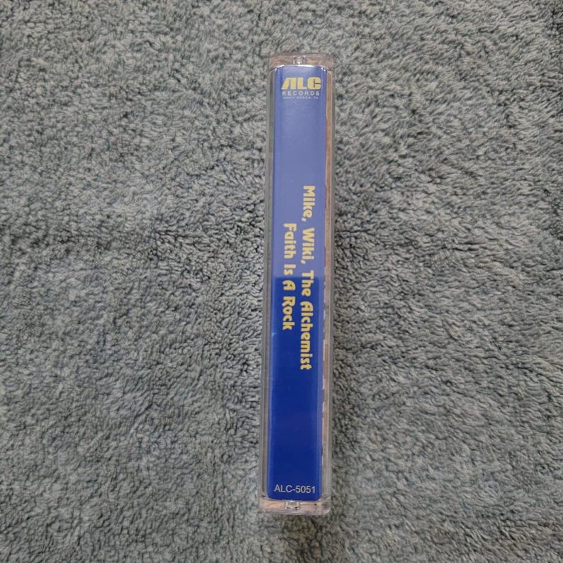May include: A blue cassette tape with the text "ALC Records" in white, "Mike, Wiki, The Alchemist" in yellow, and "Faith Is A Rock" in yellow. The cassette tape also has the text "ALC-5051" in white.