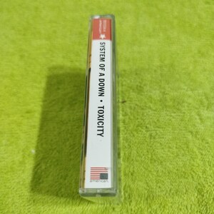 May include: A white cassette tape case with the text "SYSTEM OF A DOWN - TOXICITY" printed on the spine. The case has a red star and the word "american" printed on the bottom.