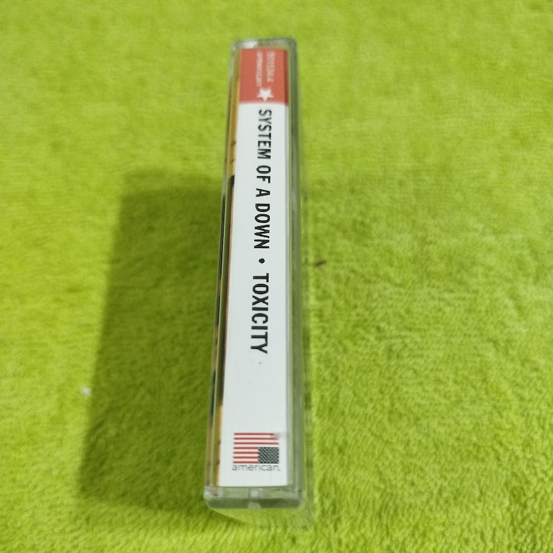 May include: A white cassette tape case with the text "SYSTEM OF A DOWN - TOXICITY" printed on the spine. The case has a red star and the word "american" printed on the bottom.