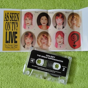 May include: A clear cassette tape labeled "Halsey The Great Impersonator" sits below a folded card with images of Halsey and the text "AS SEEN ON TV? LIVE". The card has a yellow and black color scheme.