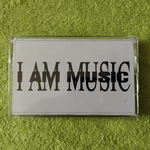 May include: A clear cassette tape case with a white insert. The insert features the text "I AM MUSIC" in a bold, black font. The background is a textured, green surface.