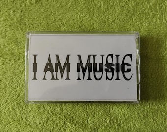 May include: A clear cassette tape case with a white insert. The insert features the text "I AM MUSIC" in a bold, black font. The background is a textured, green surface.